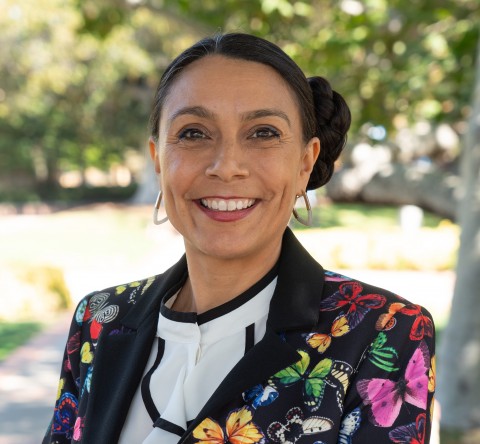 Veronica Terriquez smiling outdoors in a colorful floral blazer, with her hair pulled back and greenery softly blurred behind her.