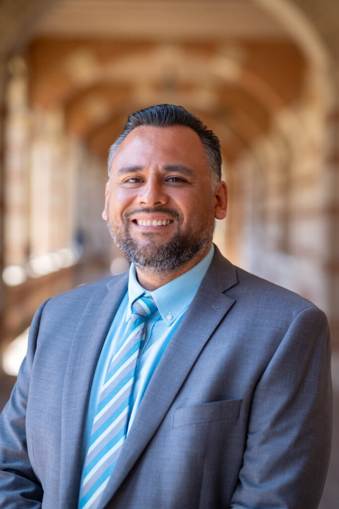 César Oyervides-Cisneros smiling in a grey suit and blue tie, with Royce Hall hallway softly blurred behind him.