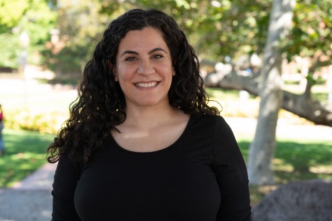 Krystell Jimenez smiling outdoors in a black top, with long curly hair and leafy green trees behind her.