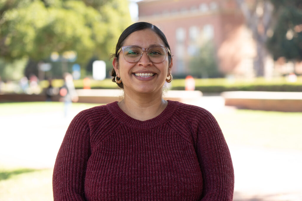 Magaly Arias smiling outdoors in a burgundy sweater and glasses, with trees and a UCLA campus building blurred behind her.