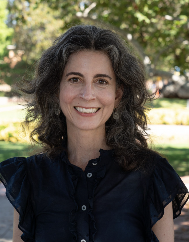 Rebecca Epstein smiling outdoors in a navy blue top, with wavy dark hair, and with leafy trees blurred behind her.