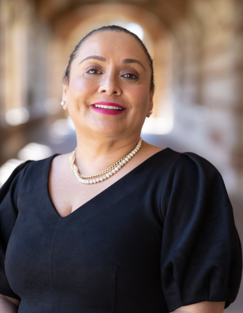 Celia Lacayo smiling in a black top and pearl necklace, with her hair pulled back and Royce Hall hallway softly blurred behind her.