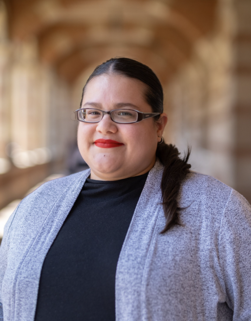 Xaviera Flores wearing a gray cardigan and glasses, with Royce Hall hallway blurred behind her.