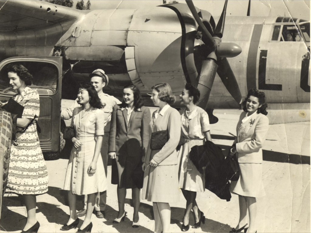 Black and white photo of seven women from the Panel of Americans standing in front of a military airplane at an unidentified base in 1945.