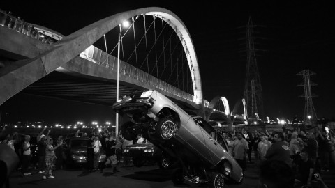 Black and white photo of a car lifted under a bridge at night, surrounded by a crowd.