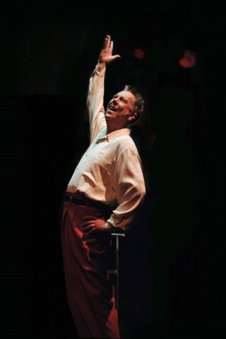 Photo of Dan Guerrero performing in a white shirt and dark pants, smiling and raising one arm under a spotlight.