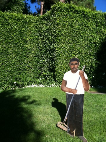 Photo of a cardboard cut-out of a man holding a rake and wearing a white shirt and installed on a green lawn.