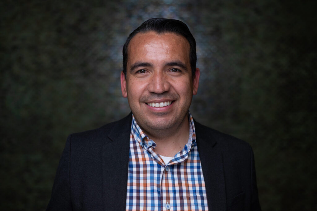 Headshot of Osvaldo Gutierrez smiling wearing a black blazer over a plaid shirt, with a black background.