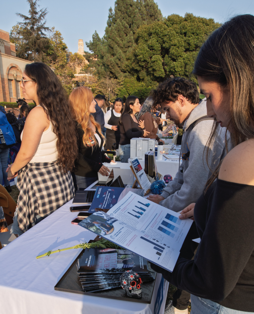 Students and attendees gathered around information tables at an outdoor UCLA event, looking at booklets.