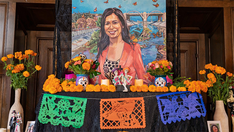 Día de los Muertos altar with orange marigolds, colorful cut-paper banners, candles, figurines, and a large painted portrait of Cindy Montañez.