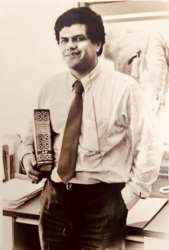 Black and white photograph of a man wearing a white shirt and tie standing beside a desk in an office.