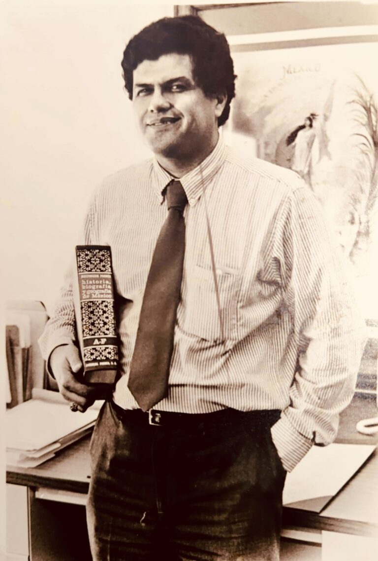 Black and white photograph of a man wearing a white shirt and tie standing beside a desk in an office.