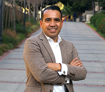 Portrait of Osvaldo Gutierrez standing outdoors at the UCLA campus, smiling with his arms crossed and wearing a brown jacket.