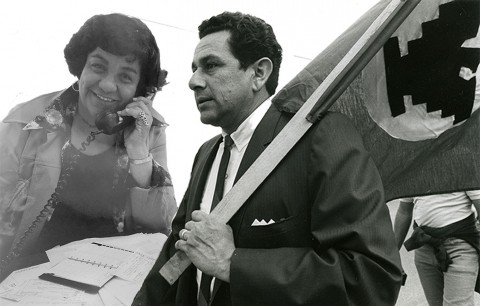 Black and white photo of a woman smiling and a man in a suit holding a flag while standing together outdoors.
