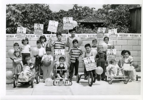 Black and white photo of children and adults posing outdoors with bicycles, balloons, and Roybal campaign signs.