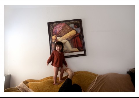 Photo of a young girl standing on a yellow couch in a living room with a framed painting hanging on the wall behind her.
