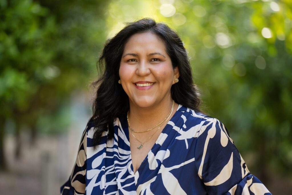 Sandra Baltazar Martínez smiling outdoors in a blue and white patterned blouse and with long dark hair, and with greenery blurred behind her.