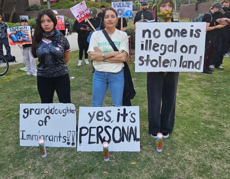 Three people standing at an outdoor demonstration holding signs supporting immigrant rights, including signs reading "granddaughter of immigrants."
