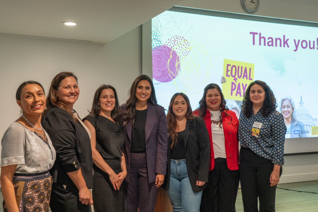Seven women posing together at a Latina Futures event in front of a presentation screen displaying an "Equal Pay" sign.