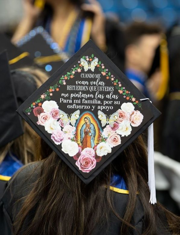 Close-up of a decorated graduation cap with colorful flowers and Spanish text, worn by a graduate at a ceremony.