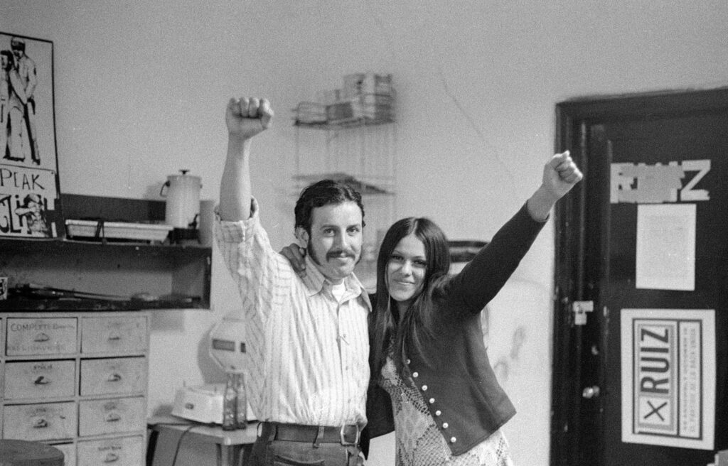 Black and white photo of a man and woman at a 1972 La Raza Unida meeting in Los Angeles, standing together with raised fists.