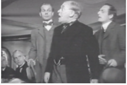 Black and white film still of five men in early 20th century suits, with two sitting, one standing and shouting, and the other two standing nearby.
