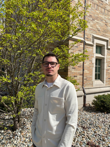 Roger Sargent Cadena, Jr. standing outdoors in front of a brick campus building and a leafy tree.