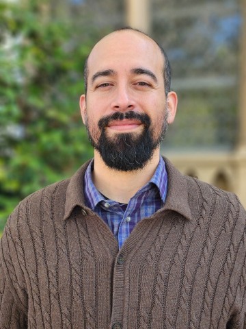 Isidro González Granados smiling outdoors in a brown sweater with greenery in the background.