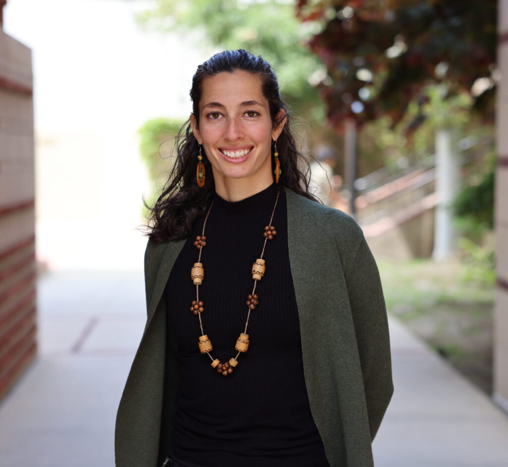 Isabella Arzeno Soltero smiling outdoors in a dark green cardigan and beaded necklace along a UCLA outdoor walkway.