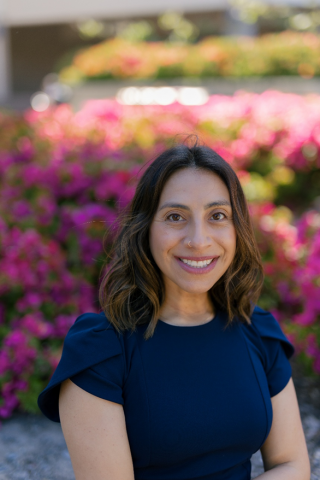 Nancy Molina-Rogers smiling outdoors in a navy blue top, with shoulder-length dark hair, and with bright pink and purple flowers blurred behind her.
