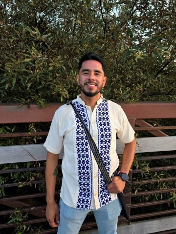 Mario Gómez Zamora smiling outdoors in a white shirt with blue embroidery, standing in front of trees and a wooden fence.