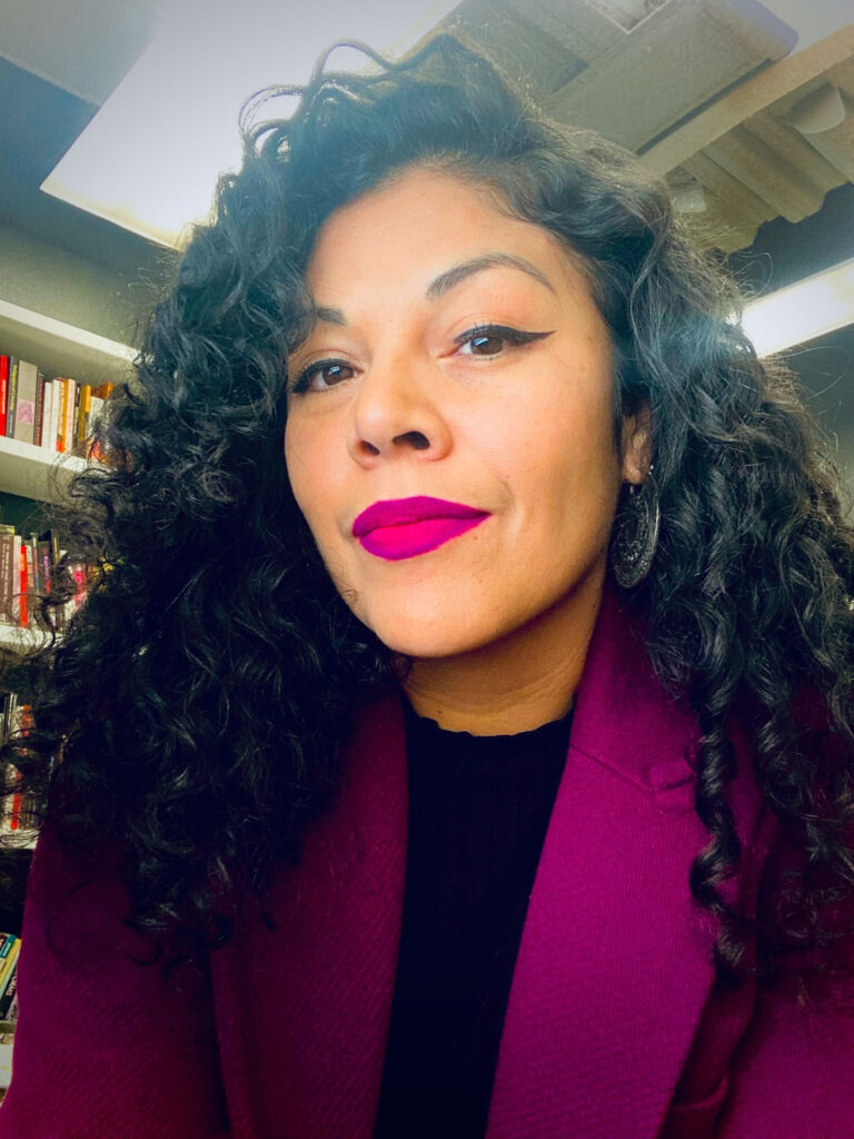 Kency Cornejo in a close-up portrait with dark curly hair with bookshelves behind her.