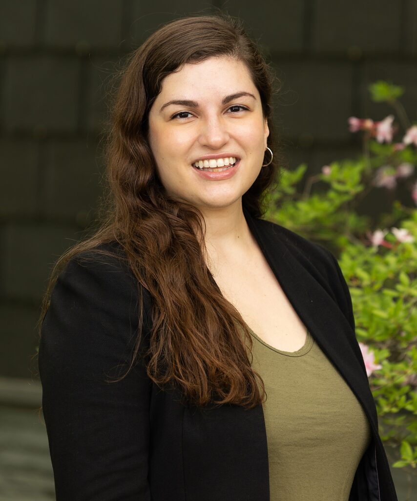 Rebecca Foote smiling outdoors in a black shirt. Leafy plants and a dark brown wall are behind her.