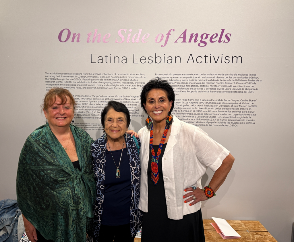 Three women posing together in front of the "On the Side of Angels: Latina Lesbian Activism" exhibition display.