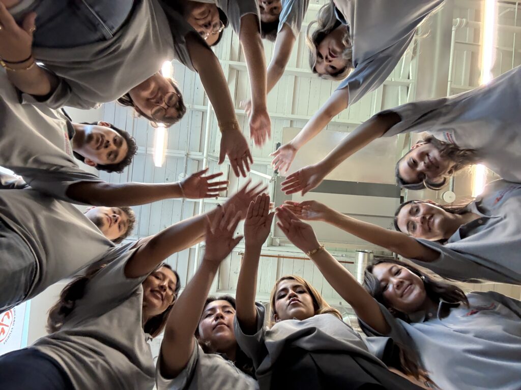 Twelve California Freedom Summer student leaders in grey shirts standing in a circle and reaching their hands toward the center at Fresno convening.