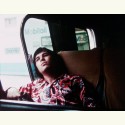 Film still of a person in a patterned shirt reclining on a bus seat while leaning against a window.