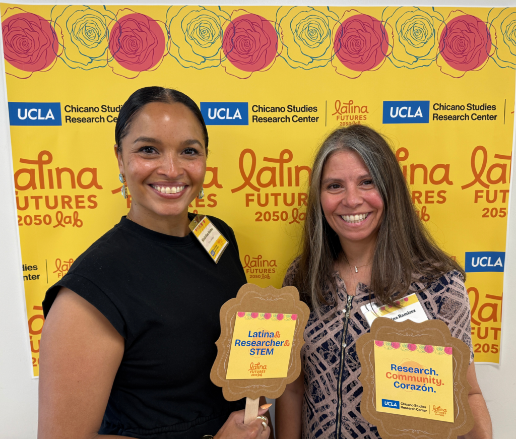 Two women smiling and holding signs in front of a step-and-repeat backdrop with Latina Futures 2050 Lab and Chicano Studies Research Center logos.
