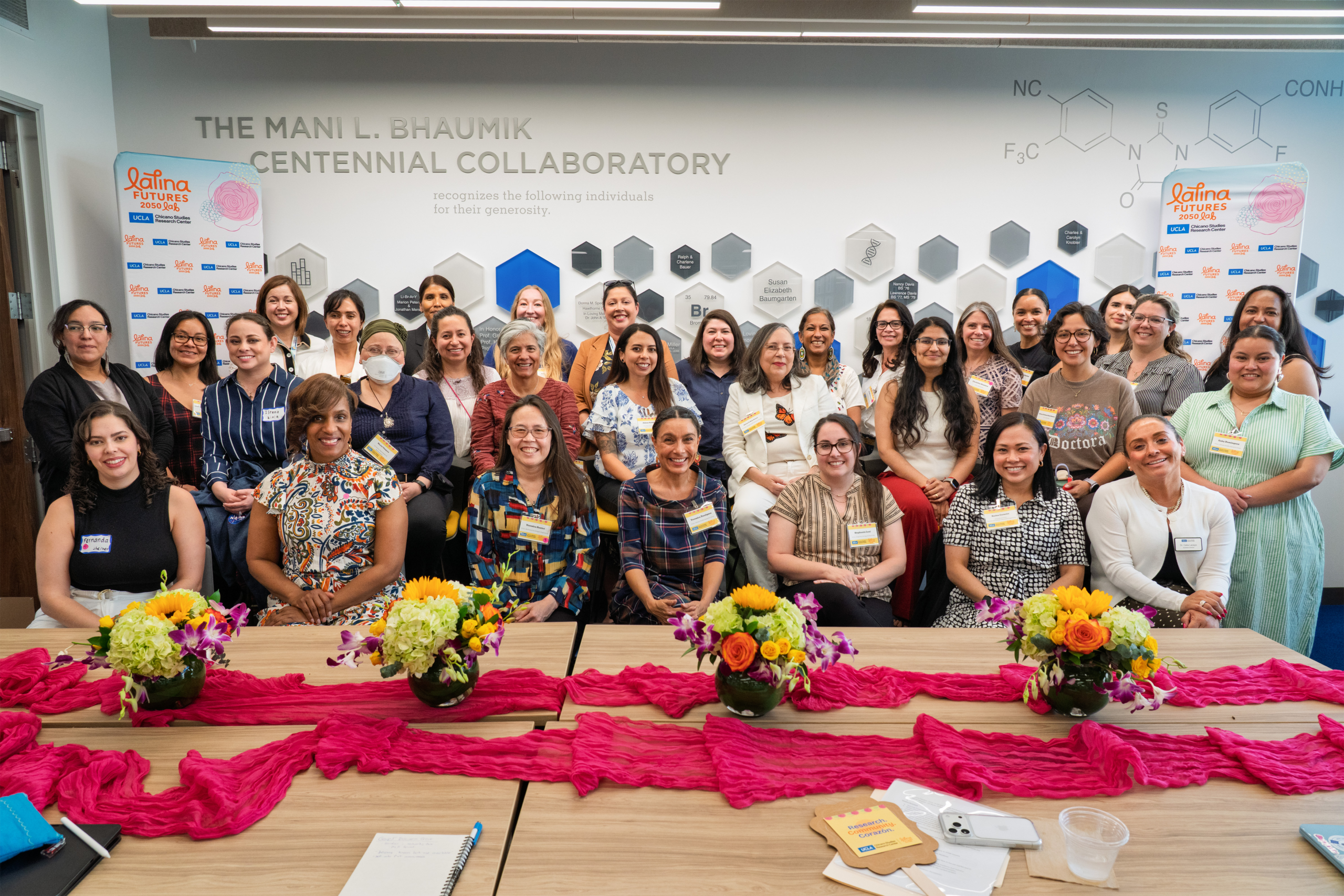 About thirty-five people posing together at a CSRC event, standing behind a table decorated with pink fabric and colorful flowers.