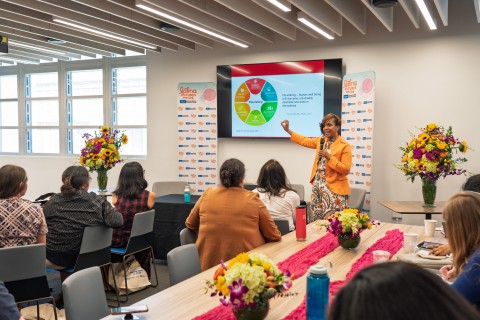 Photo of a woman in an orange jacket giving a presentation to a seated audience in a conference room decorated with colorful flowers.