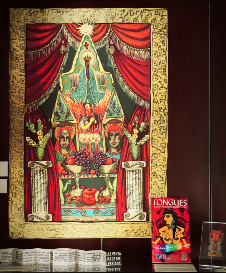 Installation photo of colorful print of red curtains, religious figures, flowers, and fruit within an illustrated frame, and a copy of Tongues zine.