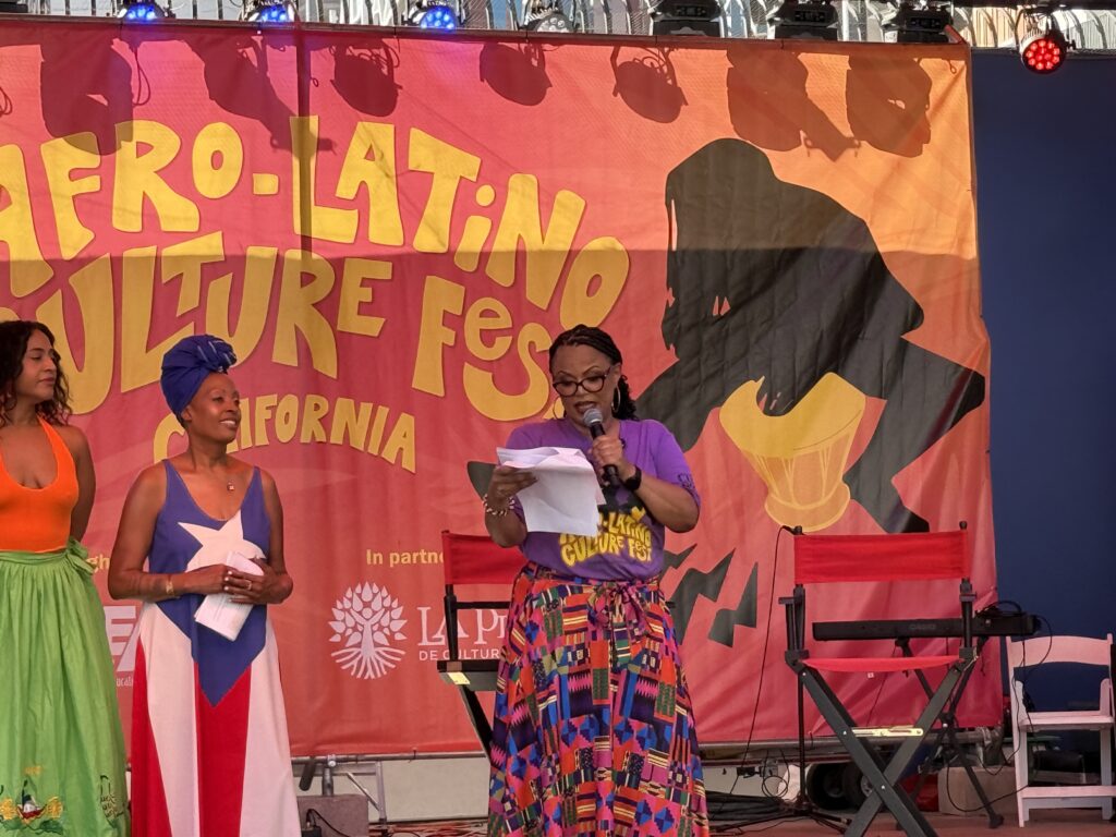 Photo of three women standing onstage, with one reading from a paper in front of a colorful Afro-Latino Culture Fest California banner.
