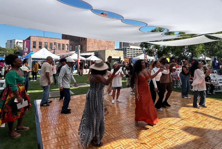 Photo of people dancing together on an outdoor dance floor at a community event, with tents and buildings, and a crowd in the background.