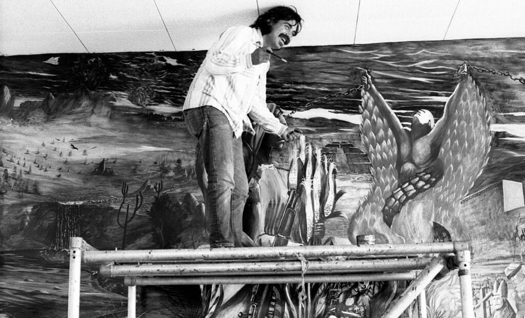 Black and white photo of Eduardo Carrillo standing on a ladder and painting the Chicano History Mural at the UCLA Chicano Research Center, c. 1970